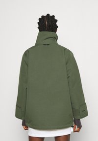 Veste vert olive, non doublée, avec un col montant, des manches larges, des accents en velcro et un cordon au bas. Elle présente un tissu texturé.