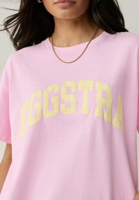 Růžové bavlněné tričko s velkým a výrazným žlutým nápisem "EGGSTRA". Kulatý výstřih, krátké rukávy a měkká textura.