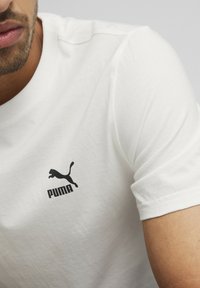 Fehér pamut póló rövid ujjakkal és kerek nyakkivágással. Az illetékes bal mellkasrészén fekete Puma logó található. Sima textúra.