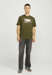 Oliven grøn kortærmet t-shirt med grafisk tryk foran. Bæres med grå løse jeans og blå sneakers med brune såler.