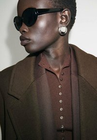 Gafas de sol oversized negras, un abrigo marrón texturizado y una camiseta de color marrón oscuro con ribetes, botones visibles y un cuello alto. Pendientes circulares de plata.