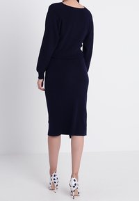 Abito a maglia blu navy con maniche lunghe e forma aderente, abbinato a tacchi bianchi con pois neri. Texture liscia e design minimalista.