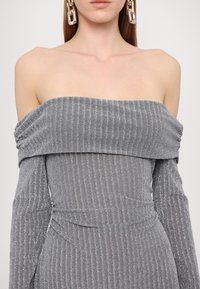 Femme portant une robe côtelée argent-gris décolletée aux épaules, manches longues, avec de grandes boucles d’oreilles métalliques et translucides liées.