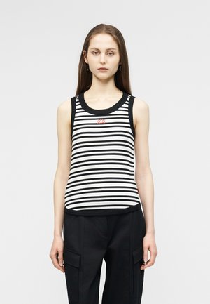 Jauna sieviete ar gariem brūniem matiem, tērpusies melnā un baltā svītraina bezroku topā un melnās cargo biksēs, stāv pret vienkāršu fonu.