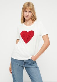 Λευκό βαμβακερό μπλουζάκι με υφασματοποιημένο κόκκινο σχέδιο καρδιάς στο μπροστινό μέρος. Η μπλούζα έχει κοντά μανίκια και στρογγυλή λαιμόκοψη.