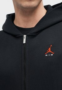 Homme portant un sweat à capuche noir zippé avec logo Jumpman rouge et texte blanc "AIR" sur la poitrine, lèvres et menton visibles.