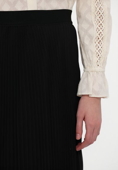 Falda negra con plisados y una cinturilla ancha; combinada con una blusa crema con encaje texturizado y puños acampanados.