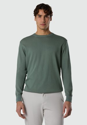 Ung mand iført en grøn rundhalset trøje med lange ærmer og lysegrå bukser, stående foran en ensartet lys baggrund.