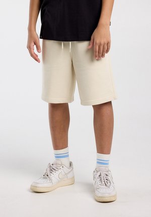 Kind draagt beige knielange korte broek, witte sneakers met blauw gestreepte sokken en een zwart shirt, en staat tegen een effen achtergrond.