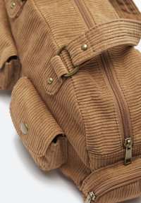 Zaino in corda marrone con tasca frontale con zip, tasca laterale con pattina e bottone a pressione, e tracolla regolabile con fibbia in ottone.