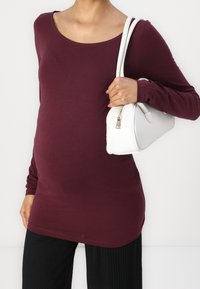 Burgundy long-sleeve top made of stretchy fabric, featuring a scoop neckline and fitted design, paired with a white handbag.