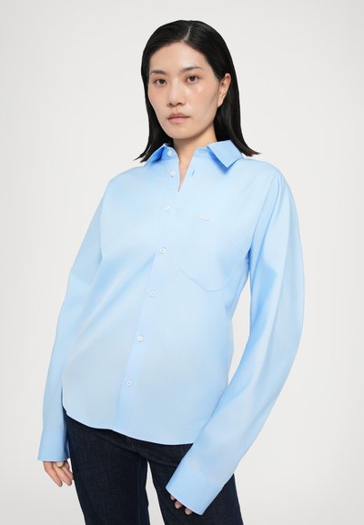 Light blue button-up shirt with long sleeves, a pocket, and a subtle logo. Made from smooth fabric, featuring a classic collar and button closures.