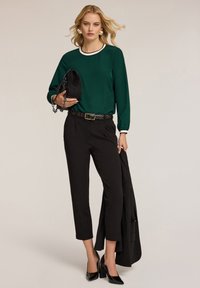 Blusa verde a maniche lunghe con bordi bianchi, pantaloni neri a vita alta, borsa nera, cintura con dettagli dorati e scarpe nere con tacco.