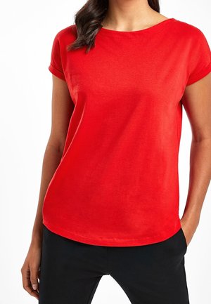 Femme portant un haut rouge vif à manches courtes avec des revers roulés et un pantalon noir, debout avec une main dans la poche et l'autre détendue.