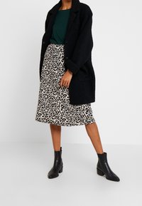 Schwarzer strukturierter Mantel, grünes Oberteil, gemusterter beiger Rock mit schwarzen Punkten und glatte schwarze Stiefeletten. Entworfen für einen gestuften Look.