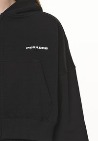 Sudadera corta con capucha negra, fabricada en material suave, con un bolsillo frontal y el logo blanco "PEGADOR" en el pecho.