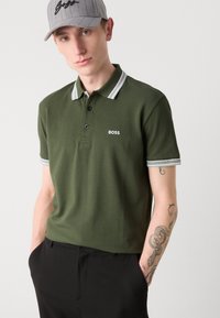 Mørkegrønn polo-skjorte med hvite detaljer på kragen og ermene, med en logo på brystet. Modellen har på seg en grå caps.