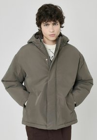 Veste matelassée vert olive avec capuche, dotée d'une fermeture éclair et de poches frontales, fabriquée en matériau résistant à l'eau, texture lisse.