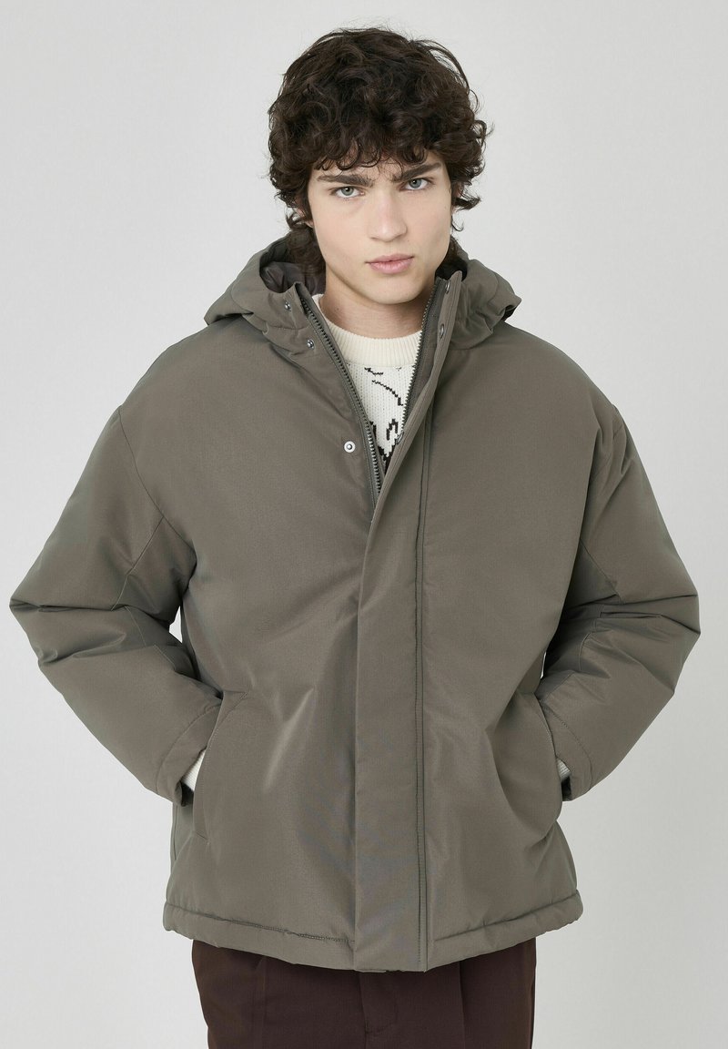 Veste matelassée vert olive avec capuche, dotée d'une fermeture éclair et de poches frontales, fabriquée en matériau résistant à l'eau, texture lisse.