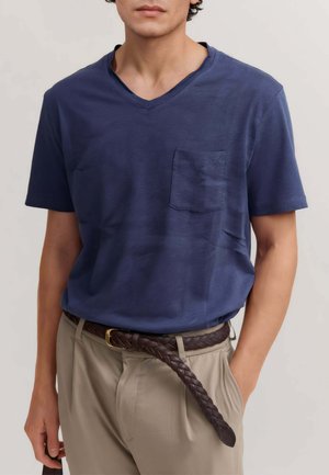 Uomo che indossa una maglietta blu navy a V con taschino sul petto e pantaloni beige con cintura marrone intrecciata che penzola sciolta.