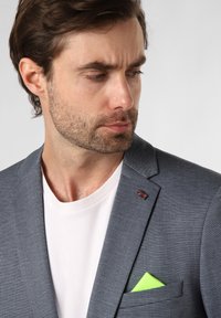 Grau karierter Blazer mit einem weißen T-Shirt darunter, ausgestattet mit einem grünen Einstecktuch und einer kleinen britischen Flaggennadel am Revers.