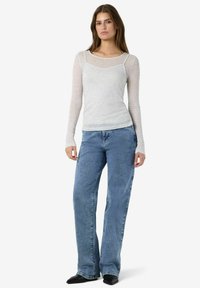 Sieviete, ģērbta caurspīdīgā baltā garajam piedurknēm augšpusē, virs balta topa, zilās augstās jostas biksēs un melnās smailējošās kurpēs, stāv.