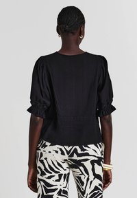 Blusa nera con maniche a palloncino in tessuto testurizzato, presenta una vita aderente e un giogo posteriore. Indossato con pantaloni bianchi che mostrano una stampa zebrate nera.