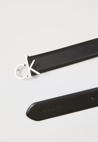 Svart lærbelte med en sølvfarget "ck" logo spenne, glatt tekstur og et rent design. Baksiden er preget med merkevaren.