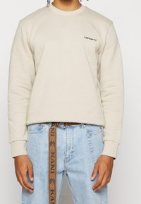 Persona vistiendo un sudadera beige de Carhartt y jeans de un azul claro, con un cinturón marrón Kani colgando de la cintura.