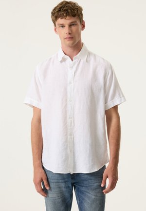 Jeune homme portant une chemise blanche à manches courtes et un jean bleu, debout devant un fond clair uni.