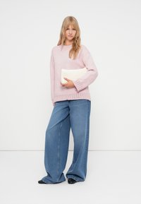 Jasnoróżowy sweter z dzianiny z prążkowanym wykończeniem, szerokie niebieskie dżinsy oraz kremowa torebka kopertówka. Modelka stoi na białym tle.