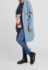 Manteau bleu clair à double boutonnage porté sur un pull bleu marine orné d'un motif de renne, associé à un jean skinny et des bottines noires à semelle marron clair.