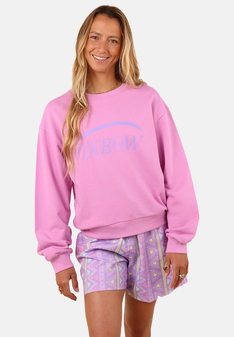 Sweatshirt rose clair avec des poignets et un ourlet côtelés, affichant "OXBOW" en lettres bleues courbées. Associé à un short pastel à motifs dans des tons lavande.