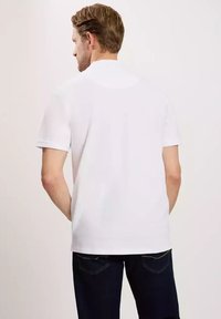 Biała krótki rękaw koszulka polo, gładki materiał, klasyczny kołnierzyk, widok z tyłu o prostym wzorze, zestawiona z ciemnoniebieskimi dżinsami.