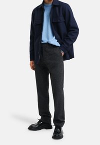 Granatowa wełniana kurtka z dwoma kieszeniami na piersi, noszona na jasnoniebieskim swetrze z dzianiny, w połączeniu z ciemnoszarymi spodniami i czarnymi skórzanymi butami.