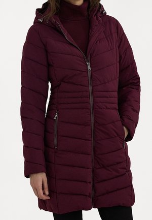Femme portant une longue veste matelassée rembourrée violet foncé avec une fermeture éclair devant, des poches latérales zippées et une capuche attachée, sur un col roulé assorti.