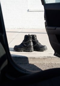 Ζευγάρι μαύρα δερμάτινα στρατιωτικά μποτάκια με χοντρές σόλες και αλυσίδες, τοποθετημένα σε πεζοδρόμιο δίπλα σε μια ανοιχτή πόρτα αυτοκινήτου.