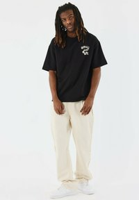 Zwarte T-shirt met korte mouwen en witte grafische afbeelding op de borst, gecombineerd met crèmekleurige wijde broek en witte sneakers.