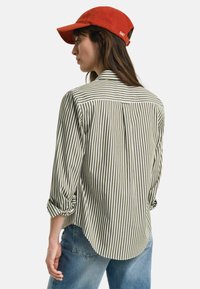 Striped shirt in olive green and white, featuring a back yoke, rolled sleeves, and a curved hem, paired with light blue jeans and a red cap.