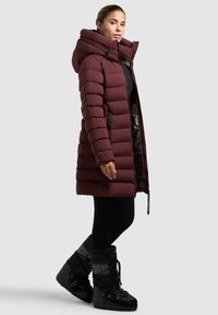 Kobieta w burgundowej pikowanej kurtce z kapturem, czarnych legginsach i czarnych zimowych butach na sznurówki stoi na tle jasnoszarym.