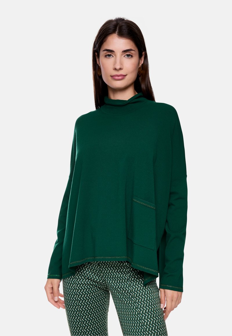 Grüner oversized Pullover mit hohem Kragen, langen Ärmeln und einer strukturierten Taschenbeschaffenheit, kombiniert mit gemusterten grünen Hosen.