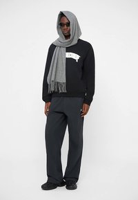 Black sweatshirt with white text, gray scarf with fringes, dark pants, and black shoes. Model wears sunglasses and a gray hood.