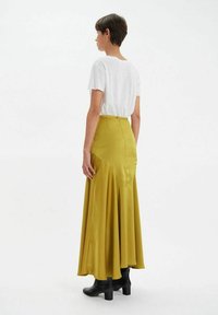 Jupe longue et fluide en satin jaune moutarde avec un ourlet évasé. Associée à un t-shirt blanc et des bottes à talons noires. Vue de dos.