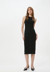 Schwarzes ärmelloses Midi-Kleid mit figurbetonter Silhouette, glatter Textur und hohem Kragen. Getragen mit schwarzen slingback High Heels mit spitzer Zehenform.