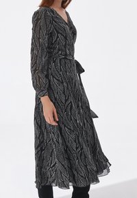 Vestido negro con patrones de hojas blancas, mangas largas, escote en V, cintura ceñida con una cinta y una falda fluida. Textura de tela semitranslúcida.