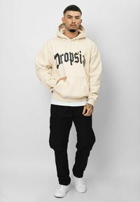 Cream hoodie with "Dropsi" graphic in black, paired with loose black cargo pants and white sneakers, featuring elastic cuffs and pockets.