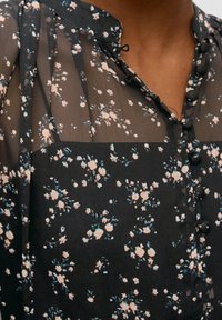 Sort gennemsigtig bluse med små rosa og hvide blomstertryk, runde stofbetrukne knapper og en høj halsudskæring.