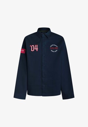 Tumši zila pogām aiztaisāma krekls ar sarkaniem un baltajiem “’04” un Red Bull Racing logotipiem krūtīs un piedurknē, garām piedurknēm, klasiskām apkaklītēm.