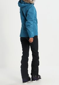 Türkisfarbene, isolierte Jacke mit einer mit Fell gefütterten Kapuze, kombiniert mit schwarzen Hosen und Schneestiefeln mit lila Akzenten. Hat ein figurbetontes Design.