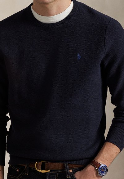 Man draagt een donkerblauwe trui over een wit overhemd, donkere jeans, een bruine riem en een polshorloge met een blauwe wijzerplaat en een bruine leren band.